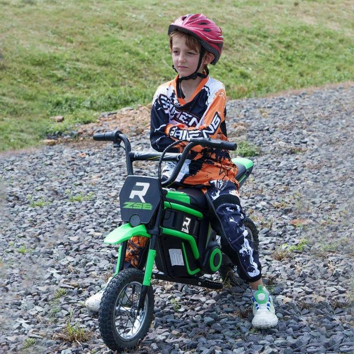 A child wearing a red helmet and motocross gear is sitting on a small, green Electric Dirt Bike with Twist Grip Throttle, Music, Horn and 12" Pneumatic Tyres 24V in Green by HOMCOM with the number "R" and "Z8E" on it. The child is on a gravel path with a grassy background.