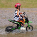 A child wearing a red helmet and motocross gear rides a green HOMCOM Electric Dirt Bike with Twist Grip Throttle, Music, Horn and 12" Pneumatic Tyres 24V through a shallow puddle on a gravel surface. The child is looking ahead while water splashes around the bike's tires.