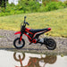 A small, red and black HOMCOM Electric Dirt Bike with Twist Grip Throttle, Music, Horn and 12" Pneumatic Tyres 24V in Red with a "3" decal on the front, parked on gravel near a grassy area. The bike is reflected in a nearby puddle of water.