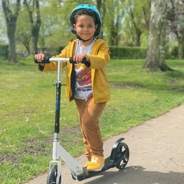 Child riding white folding kick scooter with helmet in park, adjustable handlebar visible