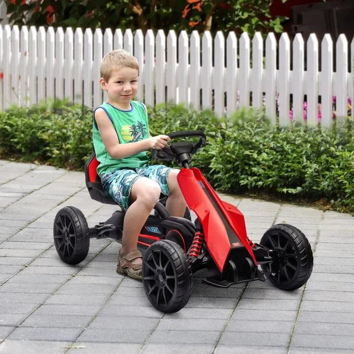Smiling boy riding red and black kids pedal go kart on outdoor path near white fence