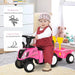 Toddler riding pink tractor ride-on toy with cargo box indoors, toy bear and dresser nearby.
