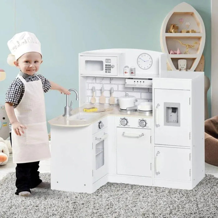 Child in chef outfit stands by white wooden kitchen playset with accessories and water dispenser.
