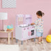 Toddler playing at pink wooden kids kitchen playset with pots, pans, and utensils in a playroom