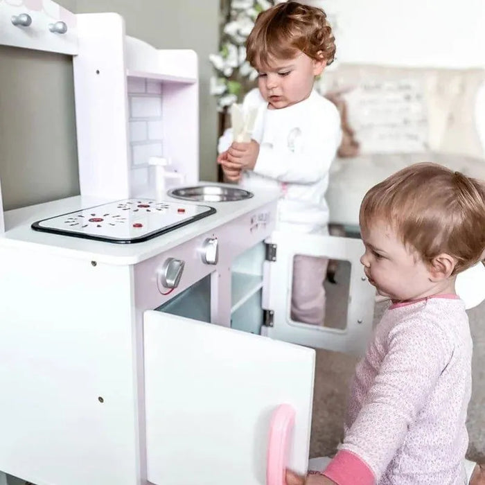 Two toddlers playing with a pink wooden kids kitchen playset and toy utensils indoors.