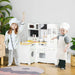 Kids in chef outfits playing with white wooden kitchen playset and accessories indoors