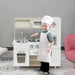 Child dressed as chef playing with white wooden toy kitchen set and stainless steel accessories