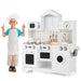 Child in chef hat and apron next to classic white wooden pretend kitchen playset with stove, oven, microwave, sink, and cooking utensils