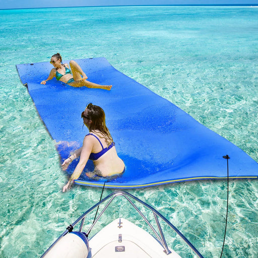 Two people relaxing on a large blue foam water float mat by a boat in clear blue water