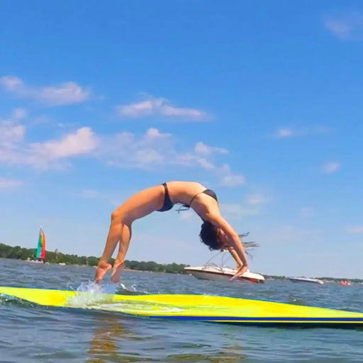 Woman in bikini doing backbend on large yellow foam water float in lake with boats