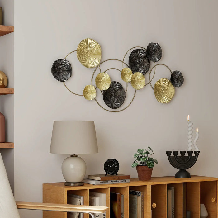 Decorative wall art with gold and black circular elements in a room with a lamp, clock, and plant on a wooden shelf.