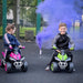 Kids riding green and pink manual ride-on quad bikes outdoors with purple smoke in background