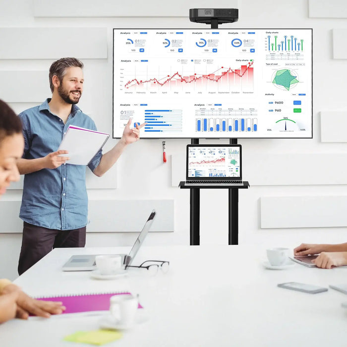 Man presenting data charts on a large TV stand with shelves and a laptop, in a modern meeting room