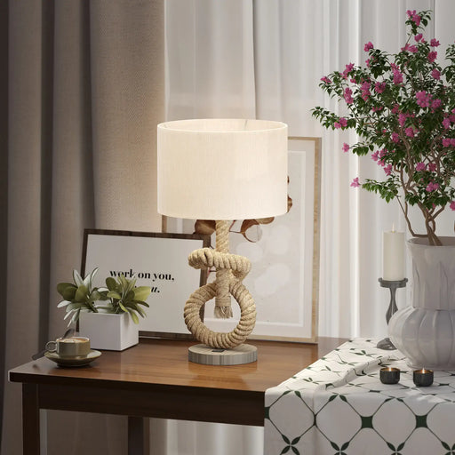 Decorative table lamp with rope design on a wooden table, surrounded by plants and candles.