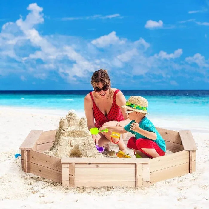 A woman and a young child build a sandcastle in an Outsunny wooden kids sandbox with cover on a sunny beach, with clear blue skies and turquoise water in the background. Both wear hats and the child uses a yellow shovel.