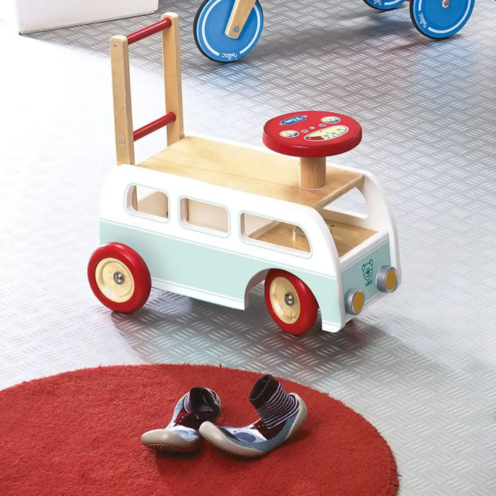Retro wooden campervan walker and ride-on toy with red wheels on a playroom floor