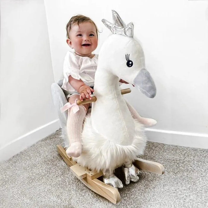 Baby on plush white and grey swan rocking horse toy with silver crown in a carpeted room