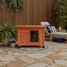 Rustic brown wooden outdoor cat house with flap door on patio, surrounded by plants and lounge chair.