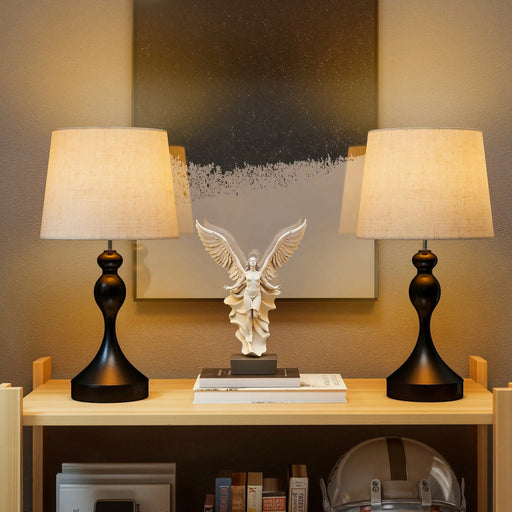 Two table lamps on a shelf with an angel statue in the center against a neutral wall.