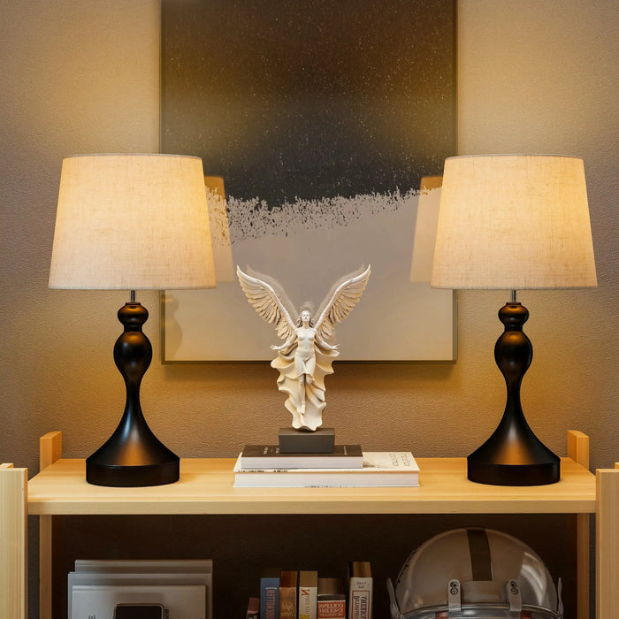 Two table lamps on a shelf with an angel statue in the center against a neutral wall.