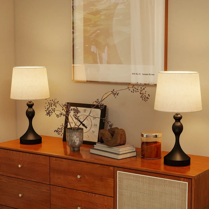 Two table lamps on a wooden dresser with decorative items and a framed picture in the background.