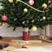 Decorated Christmas tree base with red and gold ornaments, pine cones, and beige fabric on floor
