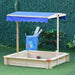 Square wooden sandbox with adjustable blue canopy in garden, toys and storage bin nearby.