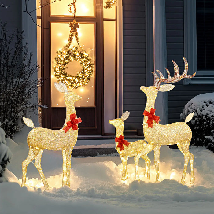 Outdoor Christmas decorations featuring three glowing reindeer with red bows in snow and a wreath-lit front door