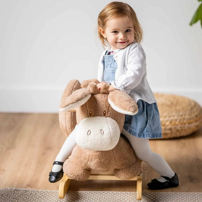 Toddler girl in white cardigan and denim dress riding a beige plush donkey rocker toy indoors