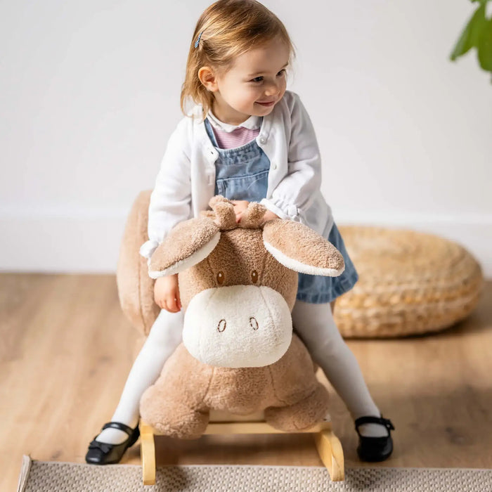 Toddler girl riding a beige plush donkey rocking toy with safety harness on wooden floor