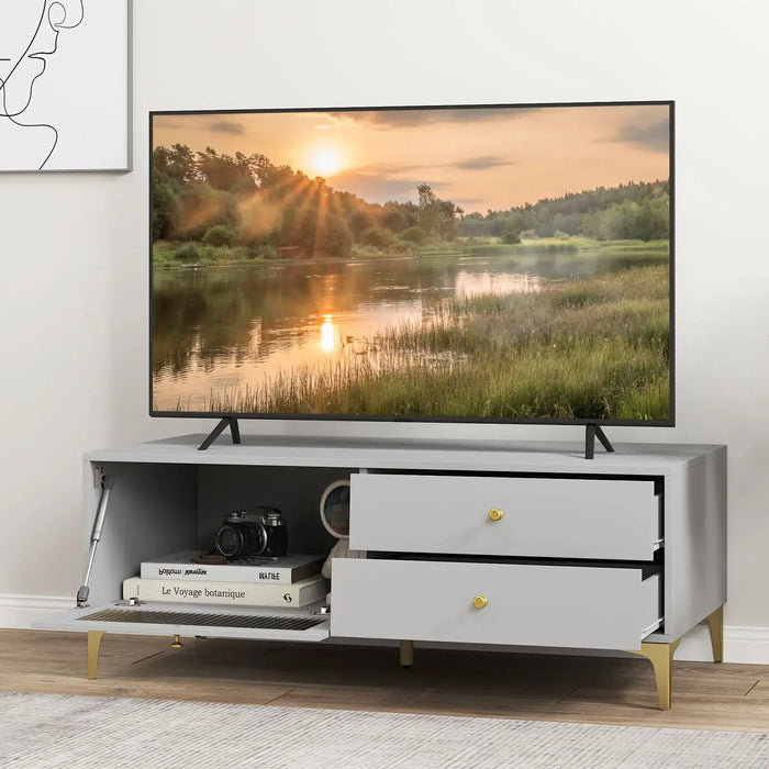 Light grey TV cabinet with open drawers and drop-down door, gold legs and knobs, holding camera and books