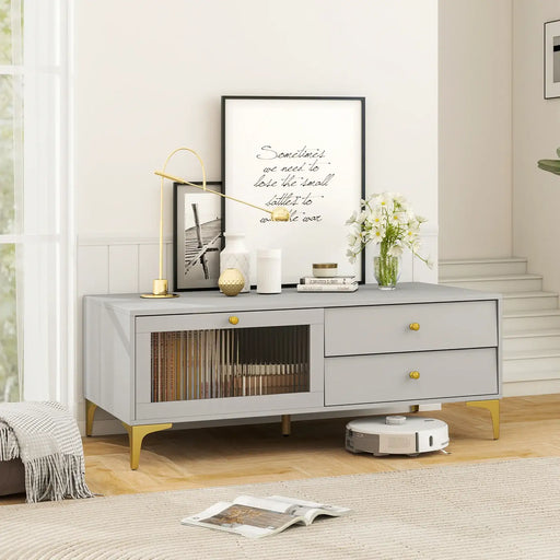 Light grey TV cabinet with gold legs and knobs, featuring drawers, drop-down door with tempered glass, decorated with lamp, vases, framed art, and flowers