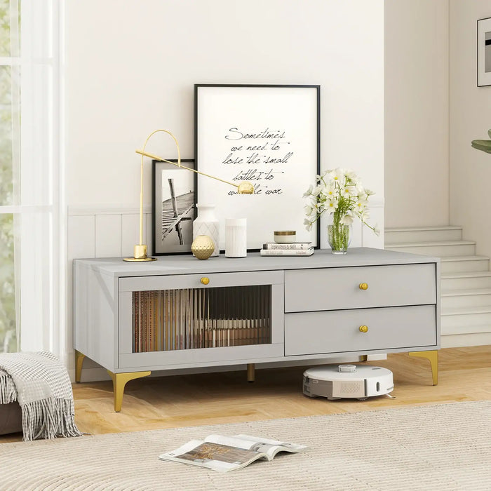 Light grey TV cabinet with gold legs and knobs, featuring drawers, drop-down door with tempered glass, decorated with lamp, vases, framed art, and flowers