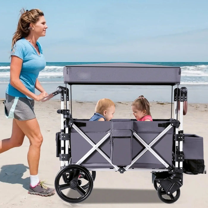 Woman pushing a gray wagon with two children on a beach.