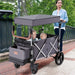 Woman pushing a gray and black folding wagon with two children on a sidewalk.