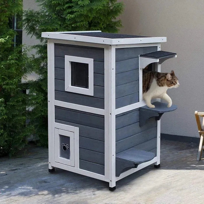 Two-tier outdoor wooden cat house in grey with white trim and steps, cat exiting side opening.