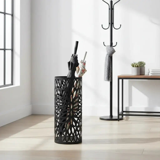 Black umbrella stand with openwork design holding an umbrella and a bag, next to a coat rack with a gray coat on a light wooden floor.