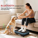 Woman exercising on a vibration plate with a dog sitting next to her in a living room.