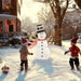 Two children in Santa hats playing in snow near a snowman with a red scarf and black top hat