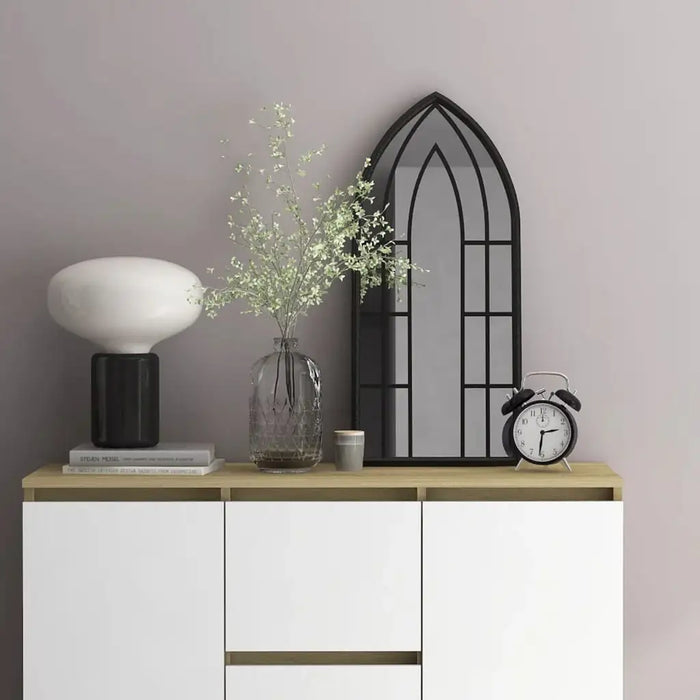 Modern wooden sideboard with white doors featuring decorative lamp, glass vase with greenery, black arched window mirror, and classic alarm clock