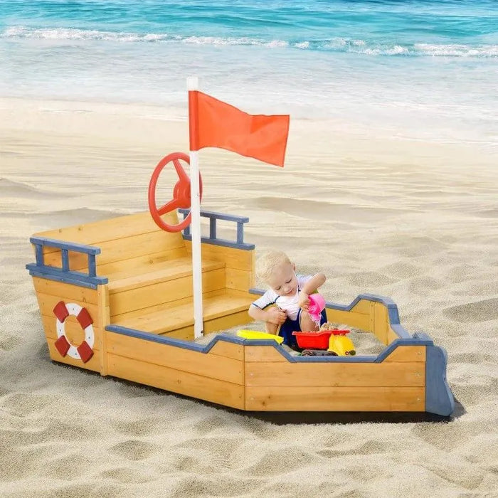 Wooden boat sandbox with red flag, steering wheel, and storage bench on sandy beach