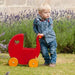 Toddler pushing cherry red wooden dolls pram toy with yellow wheels outdoors on grass