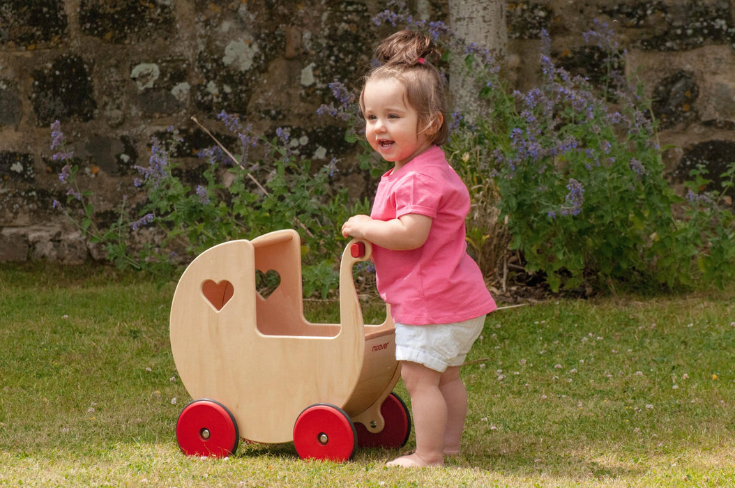 Toddler pushes natural birch wooden doll pram with red wheels outdoors on grass