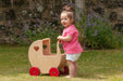 Toddler pushes natural birch wooden doll pram with red wheels outdoors on grass