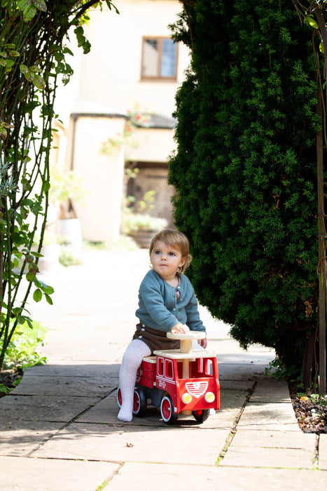 Toddler riding wooden fire truck toy outdoors, surrounded by greenery and sunlight.