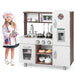 Child dressed as chef playing with wooden toddler kitchen playset featuring sink, stove, refrigerator, and accessories