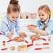 Two children playing with a wooden chef cooking set including utensils, plates, pan, and condiments.