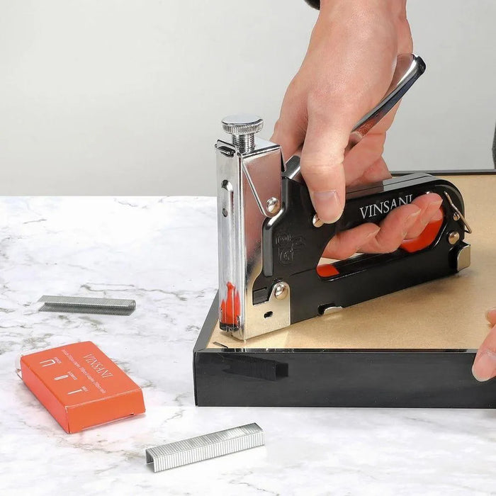 Hand using Vinsani heavy duty staple gun on wood, with box of staples on marble surface