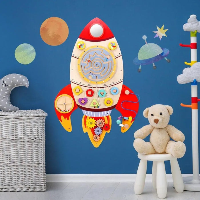 Space-themed rocket-shaped wooden activity board on blue wall in kids room with teddy bear