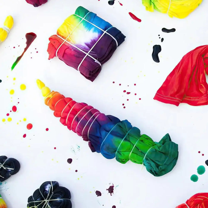Assorted tie-dye shirts and fabric wrapped in rubber bands with vibrant dye spots on white background